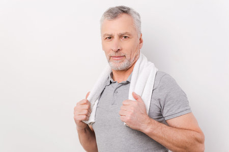 Relaxing after exercising. Confident grey hair senior man carrying towel on shoulders and looking at camera while standing against white backgroundの写真素材