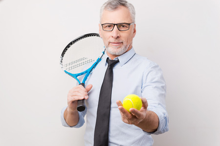 Take a break! Confident grey hair senior man in shirt and tie holding tennis racket and stretching out tennis ball while standing against white backgroundの写真素材