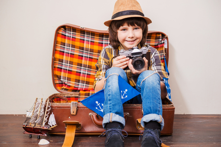 Say cheese! Little boy in headwear holding camera and smiling while sitting in suitcase against brown backgroundの写真素材