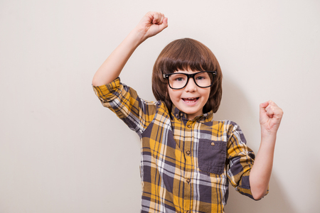 Little things can make him happy! Little boy in eyewear keeping arms raised and smiling while standing against grey backgroundの写真素材
