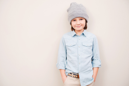 So young and so stylish! Handsome little boy in hat holding hands in pockets and looking at camera while standing against grey backgroundの写真素材