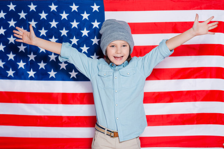 Hello America! Little boy in eyewear keeping arms raised and smiling while standing against American flagの写真素材