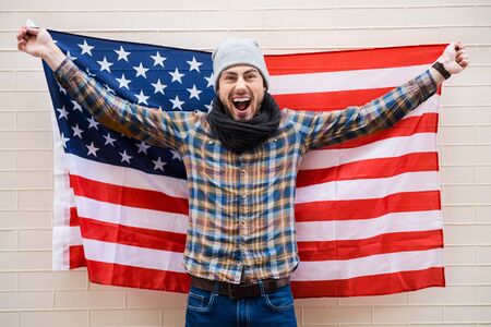 Excited patriot of American style. Excited young man holding American flag while standing against brick wallの写真素材