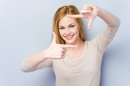 Gesturing finger frame. Beautiful young woman looking at camera and gesturing finger frame while standing against grey backgroundの写真素材