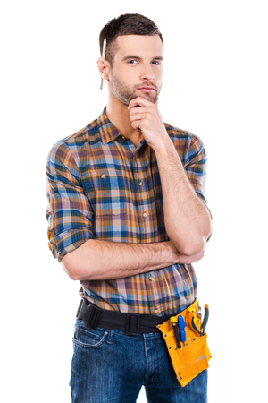 Thinking about your problem. Handsome young male carpenter with tool belt holding hand on chin and looking at camera while standing against white backgroundの写真素材