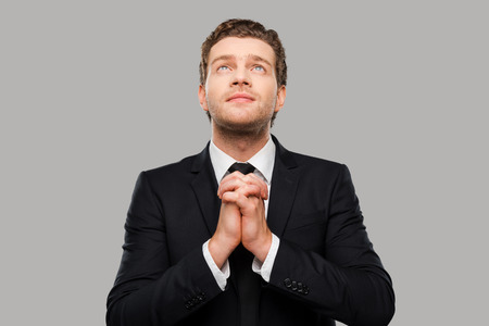 Businessman praying. Thoughtful young man in formalwear holding hands clasped and fingers crossed while standing against grey backgroundの写真素材