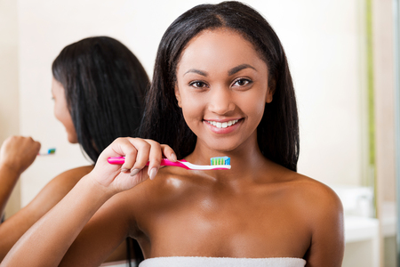 Teeth care. Beautiful young African woman brushing her teeth and smiling while standing against a mirror in bathroomの写真素材