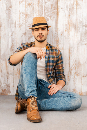 Calm and confident. Handsome young man wearing hat and looking at camera while sitting against the wooden wallの写真素材