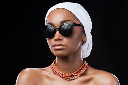 Combining fashion and traditions. Beautiful African woman wearing a headscarf and sunglasses while standing against black backgroundの写真素材