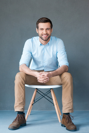 Positive thinker. Full length of handsome young man looking at camera and smiling while sitting against grey backgroundの写真素材