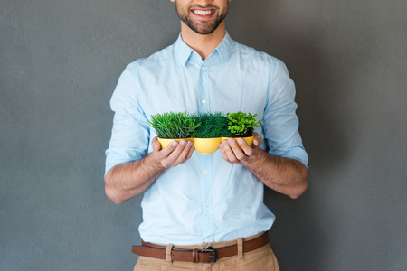 Grow up your business. Cropped picture of cheerful young man in shirt holding flower pot and smiling at camera while standing against grey backgroundの写真素材