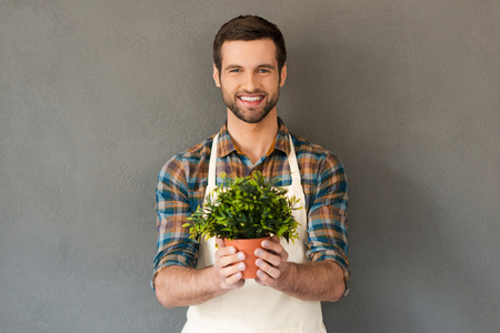 Cheerful gardener. Cheerful young gardener holding flower pot and smiling at camera while standing against grey backgroundの写真素材