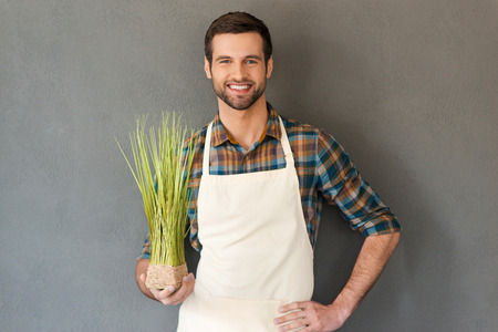 Gardening is my passion. Cheerful young gardener holding flower pot and smiling at camera while standing against grey backgroundの写真素材