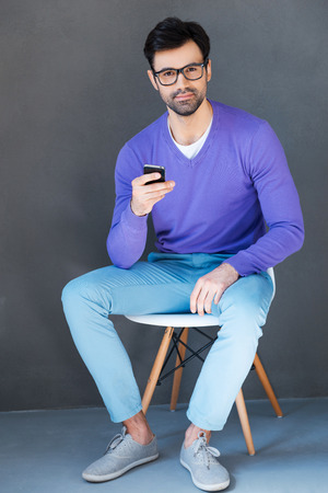 Young and creative. Handsome young man holding mobile phone and looking at camera while sitting against grey backgroundの写真素材