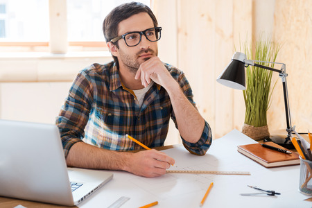 Waiting for inspiration. Thoughtful young man holding hand on chin and looking away while sitting at his working place Waiting for inspiration. Thoughtful young man holding hand on chin and looking away while sitting at his working place