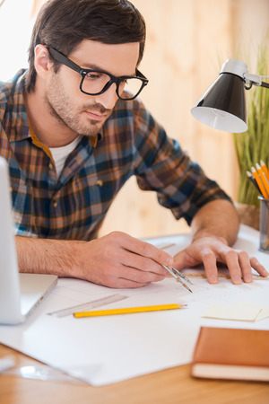 Turning his ideas to reality. Concentrated young man using compass for drawing while sitting at his workplaceの写真素材