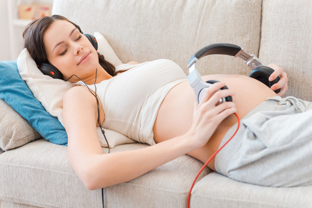 We love music. Beautiful pregnant woman holding headphones on her belly while lying on sofaの写真素材