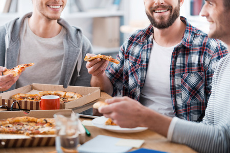 Pizza for productive work. Cropped image of three happy young men eating pizza while sitting at the desk together Pizza for productive work. Cropped image of three happy young men eating pizza while sitting at the desk together