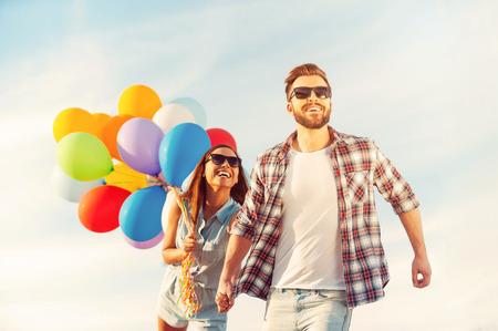 Living life to the fullest. Cheerful young couple holding hands and smiling while walking outdoors with colorful balloonsの写真素材