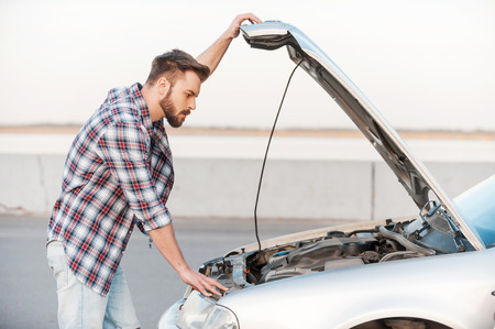 Car trouble. Serious young man holding hands on vehicle hood and looking inside it while standing outdoorsの写真素材