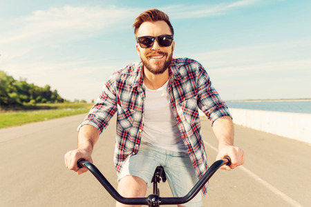 Life is brighter when you are on bike. Handsome young man in eyewear smiling at camera while riding on bicycle along the roadの写真素材