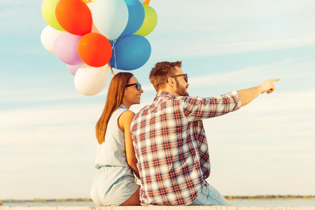 This view is stunning! Smiling young couple looking away while sitting on parapet with colorful balloonsの写真素材