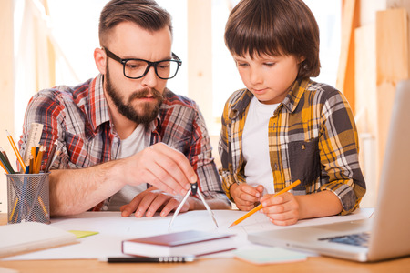 Turning their dreams to reality. Concentrated young man using compass for drawing while sitting at his workplace with his sonの写真素材