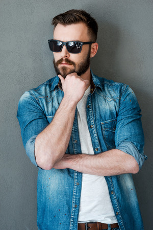Casually handsome. Thoughtful young man holding hand on chin and looking away while standing against grey backgroundの写真素材