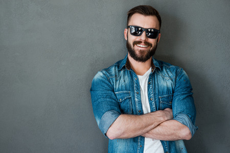 Looking handsome and casual. Happy young man keeping arms crossed and smiling at camera while standing against grey backgroundの写真素材