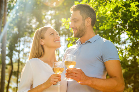 Low angle view of happy young loving couple holding glasses with white wine and looking at each other with smile while standing outdoors togetherの写真素材