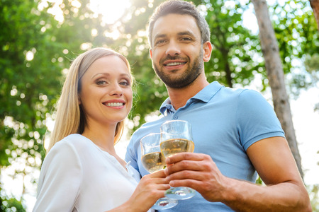 Low angle view of happy young loving couple holding glasses with white wine and smiling while standing close to each other outdoorsの写真素材