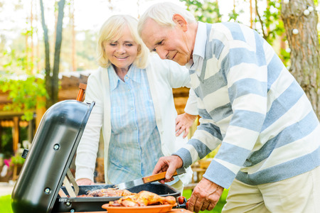 Happy senior couple barbecuing meat on the grill while standing at the back yard of their houseの写真素材