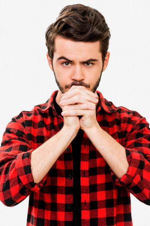 Hoping for the best. Thoughtful young man holding hands clasped near face and looking at camera while standing against white backgroundの写真素材
