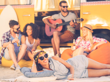 Summer time with friends. Cheerful young woman smiling at camera while lying near retro minivan with friends in the backgroundの写真素材