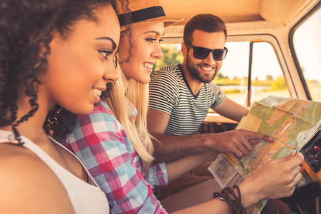 Planning their road trip. Side view of three cheerful young people examining map and smiling while sitting inside of their minivanの写真素材