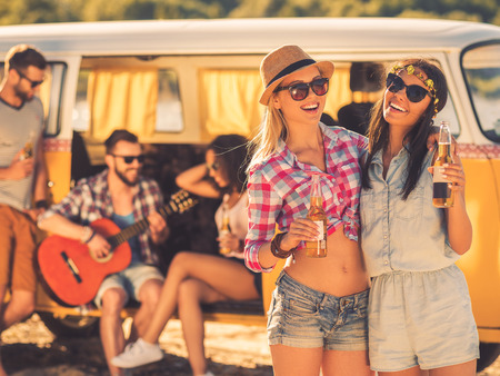 Always together and never apart. Two cheerful young women holding bottles of beer while their friends sitting at the background in retro minivanの写真素材