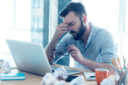 Feeling exhausted. Frustrated young beard man massaging his nose and keeping eyes closed while sitting at his working place in office Feeling exhausted. Frustrated young beard man massaging his nose and keeping eyes closed while sitting at his working place in office