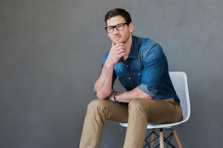Confident young man. Thoughtful young man holding hand on chin while sitting against grey background