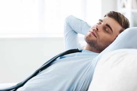 Time for relaxation. Handsome young businessman holding hands behind head and keeping eyes closed while sitting on sofaの写真素材