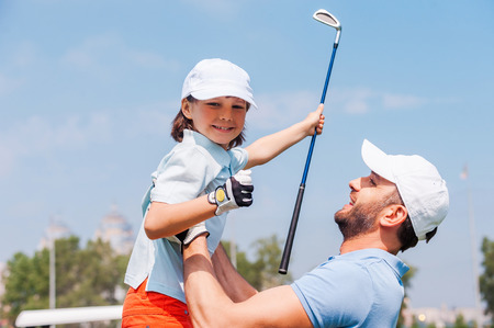 Little winner. Cheerful young man picking up his son while standing on the golf courseの写真素材
