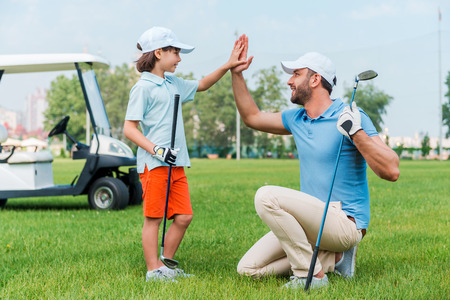 My little winner! Cheerful young man and his son giving high-five to each other while standing on the golf courseの写真素材