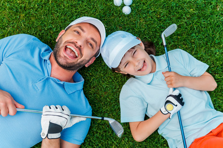 Happy golfers. Top view of cheerful little boy and his father holding golf clubs and smiling while lying on the green grassの写真素材