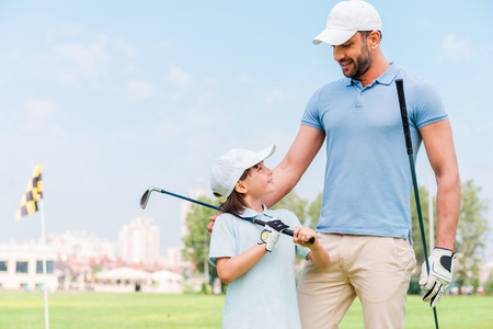 Little golfer with father. Happy young man and his son holding golf clubs and looking at each other while standing on the golf courseの写真素材