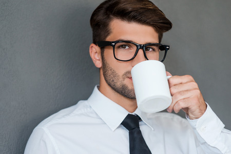 Inspired with cup of fresh coffee. Confident young man in shirt and tie drinking coffee and looking away while standing against grey backgroundの写真素材