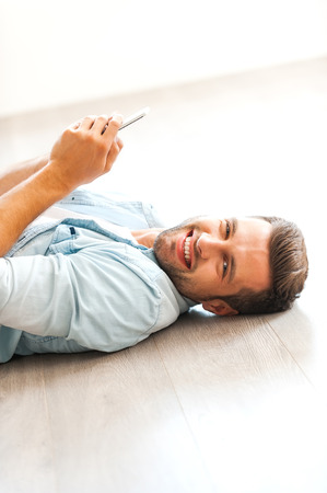 Relaxing at home. Cheerful young man holding mobile phone and looking at camera while lying on the floor at his apartmentの写真素材