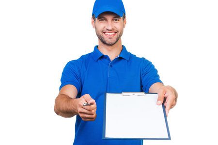 Sign please! Smiling young courier stretching out clipboard and pen for signing while standing against white backgroundの写真素材