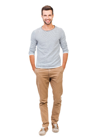 Confident and stylish. Full length of cheerful young man holding hands in pockets and looking at camera while standing against white background Confident and stylish. Full length of cheerful young man holding hands in pockets and looking at camera while standing against white background