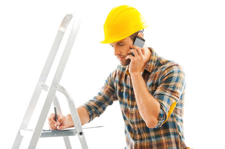 Construction is a serious business. Serious young repairman writing in clipboard and talking on the...