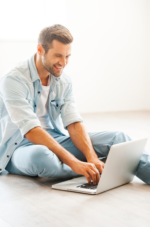 Being at home in pleasure. Handsome young man working on laptop and smiling while sitting on the floor at his apartmentの写真素材