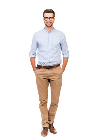 Confident in any situation. Happy young man holding hands in pockets and looking at camera while standing against white background